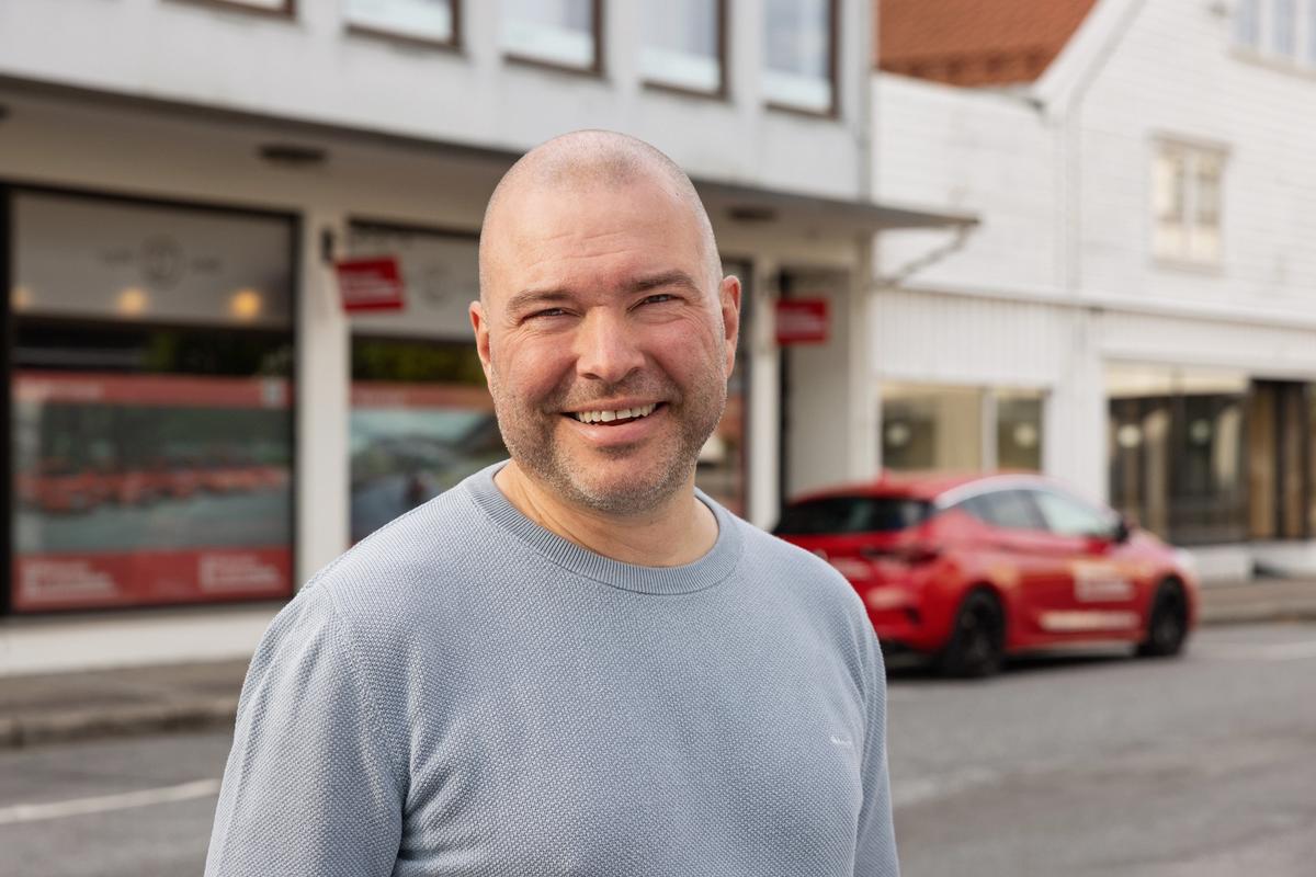Mann med lys genser smiler i kamera. I bakgrunnen ser vi en rød kjøreskolebil.