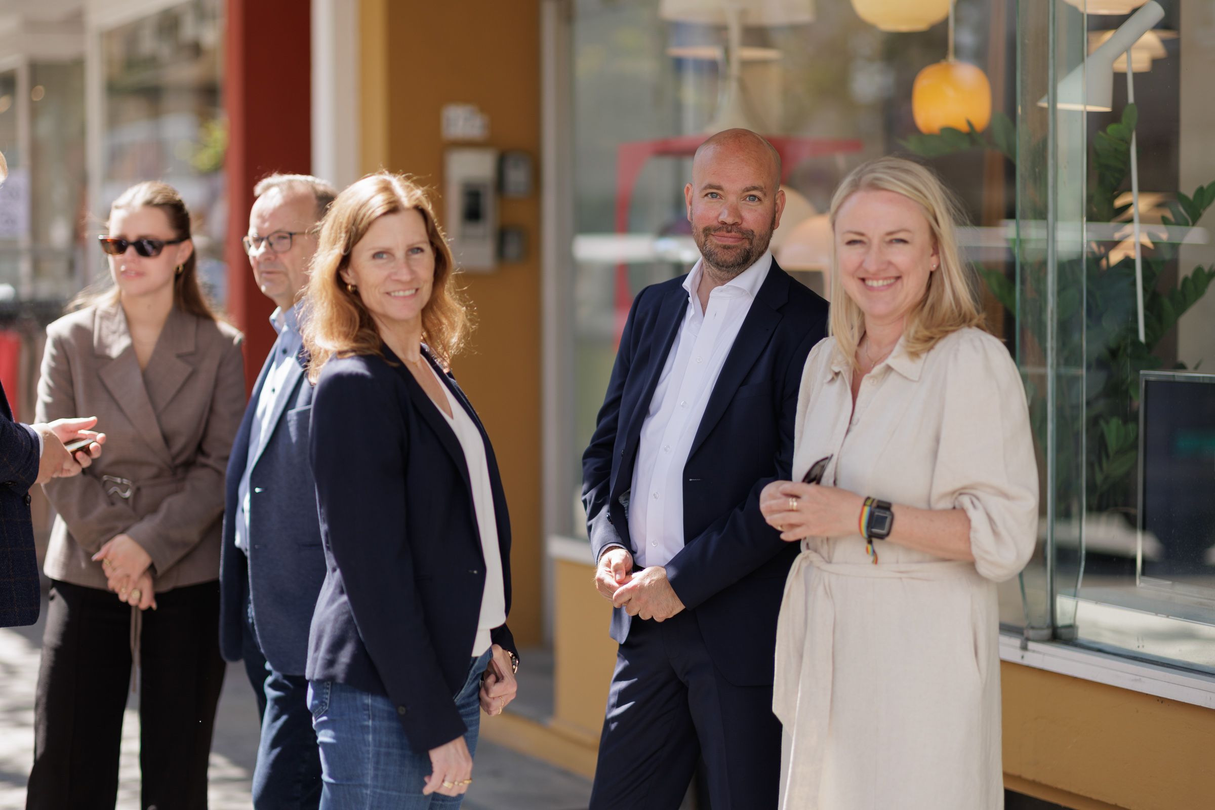 Bildet viser tre glade mennesker i solskinn i en handlegate som smiler mot kamera.