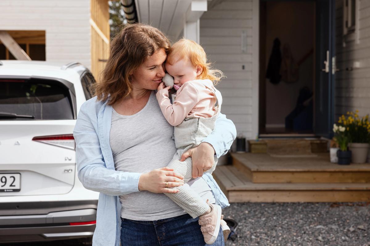 Gravid dame som holder en liten jente. Hus og bil i bakgrunnen.
