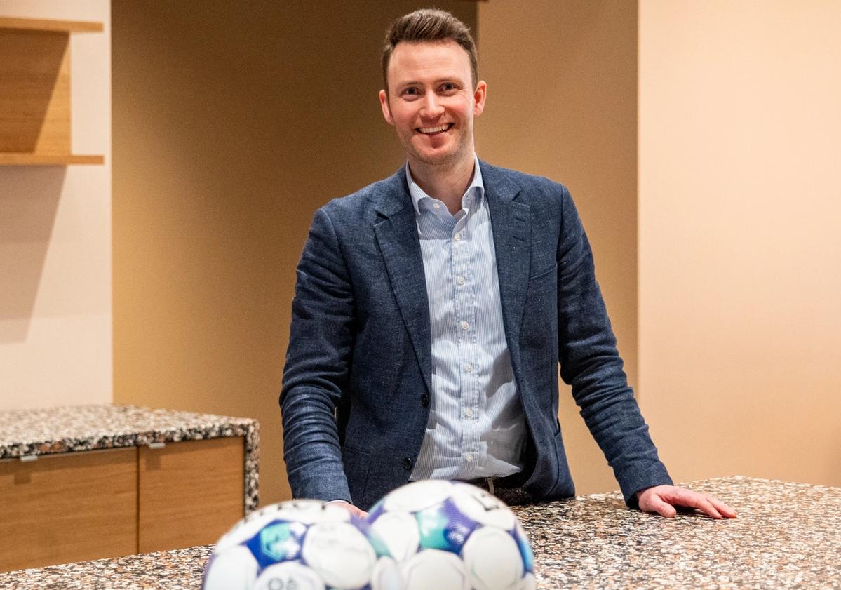 Mann med dress står ved en kjøkkenbenk med hendene på platen. I front av bildet ligger to fotballer.