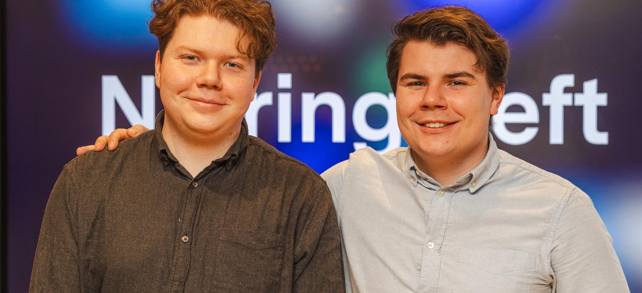 To menn med skjorte står sammen og smiler i kamera. Mann til høyre holder hånden på skulderen til den andre. I bakgrunn en skjerm der man skimter at det står Næringsteft.