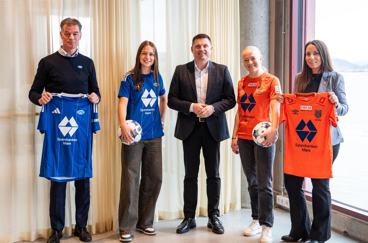 Fem mennesker står på rekke og smiler i kamera. To kvinner har fotballdrakt på og holder en ball. To holder drakter i hendene.