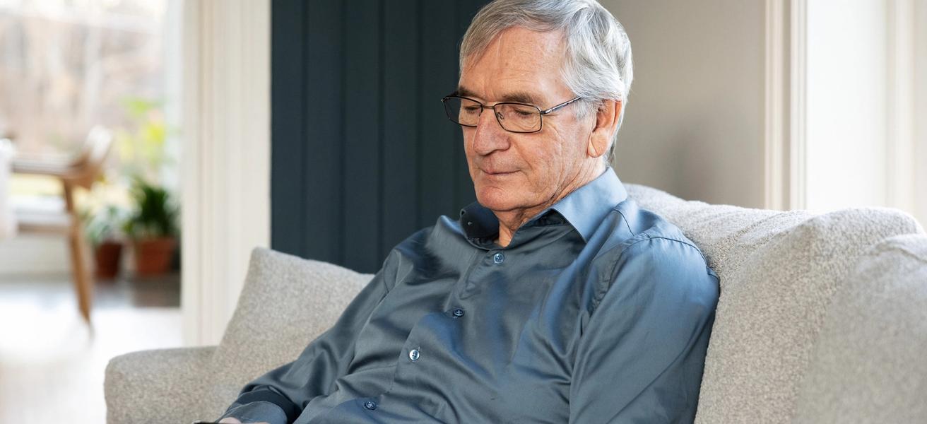 Eldre mann med blå skjorte, sitter i sofa og ser i mobiltelefonen.