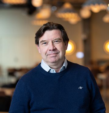Mann med mørkt hår, mørk genser med skjorte under. Smiler og ser i kamera. Blurret bakgrunn