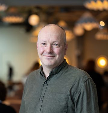 Mann med mørk grønn skjorte, smiler og ser i kamera. Blurret bakgrunn.