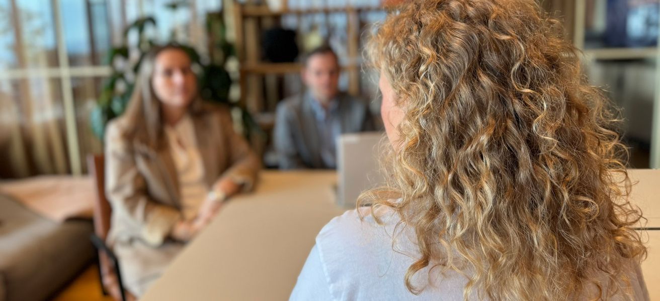 Bildet viser tre personer rundt et bord. I front er det en kvinne med blondt hår som sitter med ryggen mot kamera. På andre siden av bordet sitter en mann og en kvinne. De har blikket på kvinnen i front.