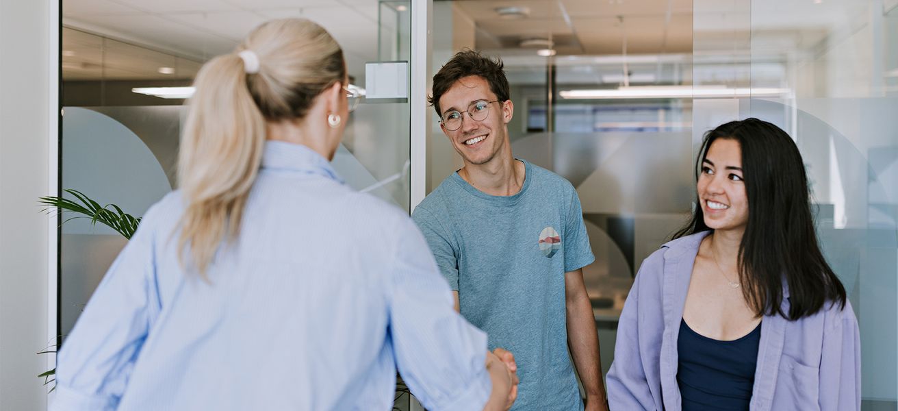 To unge mennesker i et kontorlokale hilser på dame med ryggen til.