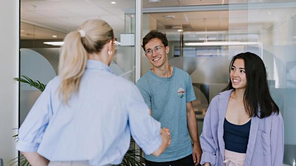 To unge mennesker i et kontorlokale hilser på dame med ryggen til.