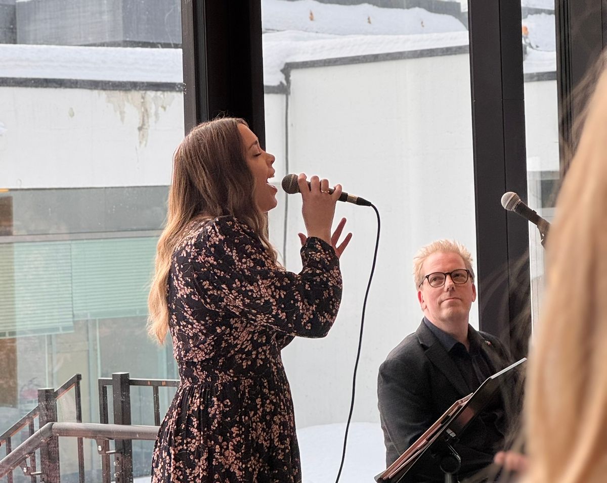 Dame sett fra siden som holder en mikrofon og synger. Mann i bakgrunnen.