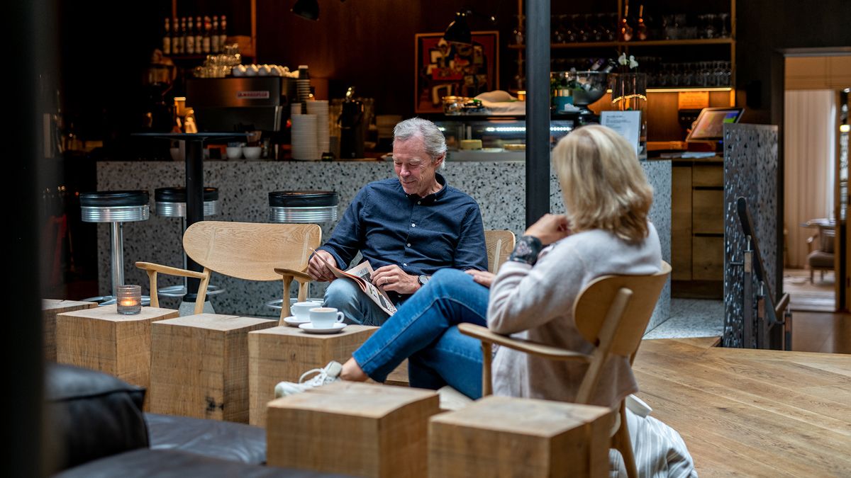 Mann og dame sitter på kafe. Damen sitter med ryggen til, mannen leser en avis. På bordet foran står det to hvite kaffekopper.