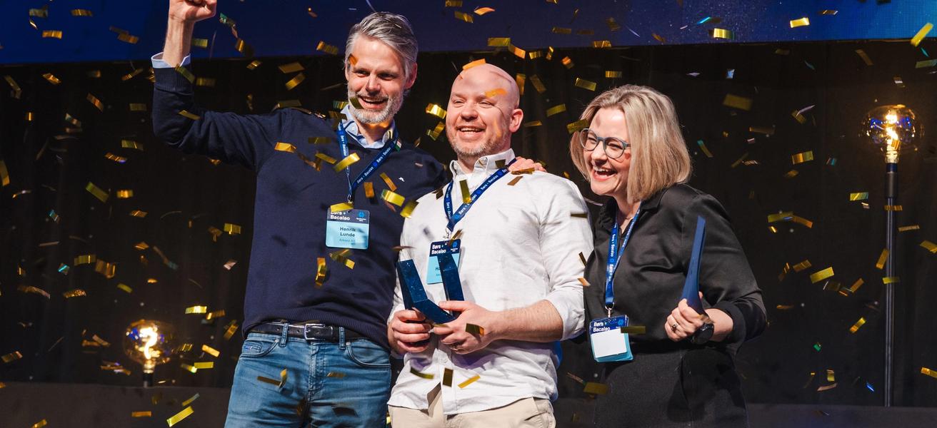 Flere som holder en pokal og jubler, på en scene. Konfetti og god stemning.