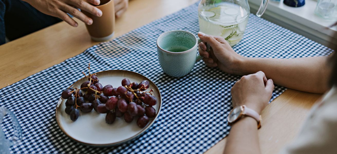 To personer sitter ved et kjøkkenbord, det er kun armene som er synlig. De holder hver sin kaffekopp og det står et fat med røde druer på bordet.