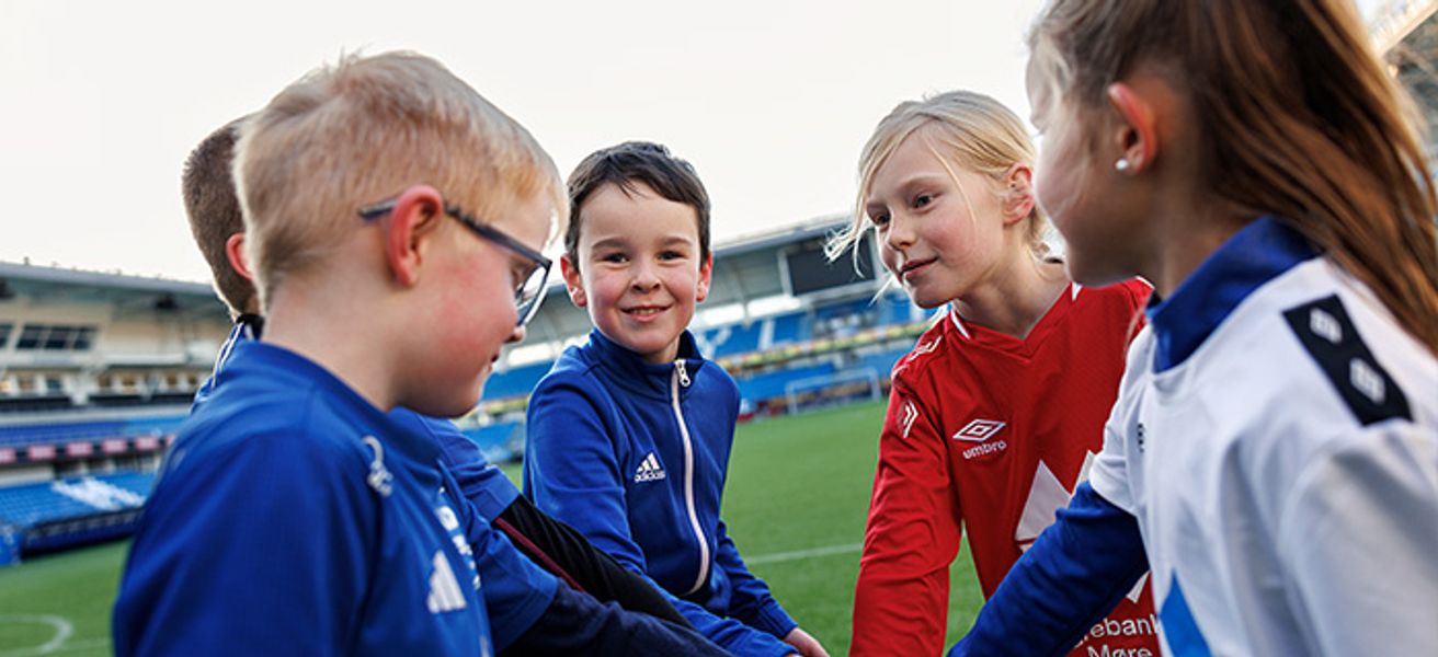 Gutter og jenter som står i en ring med hendene inn i midten, på en fotballbane. En gutt ser i kamera.