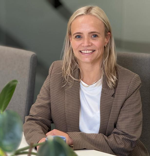 En dame med blondt hår og dressjakke sitter ved et bord og smiler til kamera