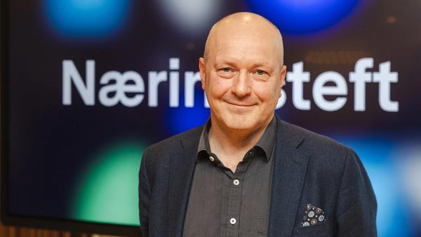 Mann uten hår, med dressjakke og skjorte ser i kamera. Skjerm med Næringsteft bak.