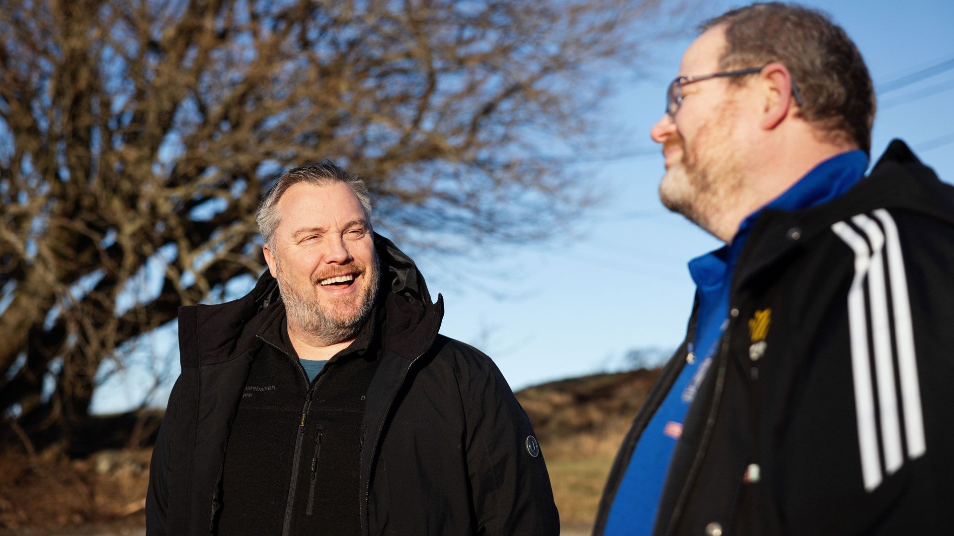 To menn står ute og prate. I bakgrunnen er et stort tre. Den ene mannen ser på den andre og smiler.