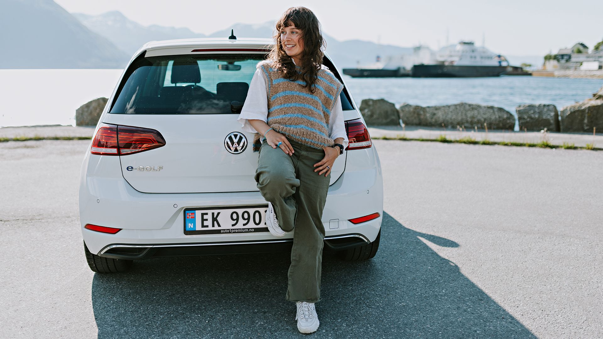 Ung dame som lener seg til bagasjerommet på en hvit WV-bil. Ferge i bakgrunnen. 