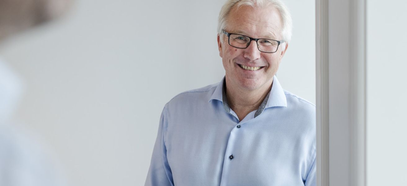 Mann i blå skjorte lener seg mot vinduskarm, ser i kamera og smiler.