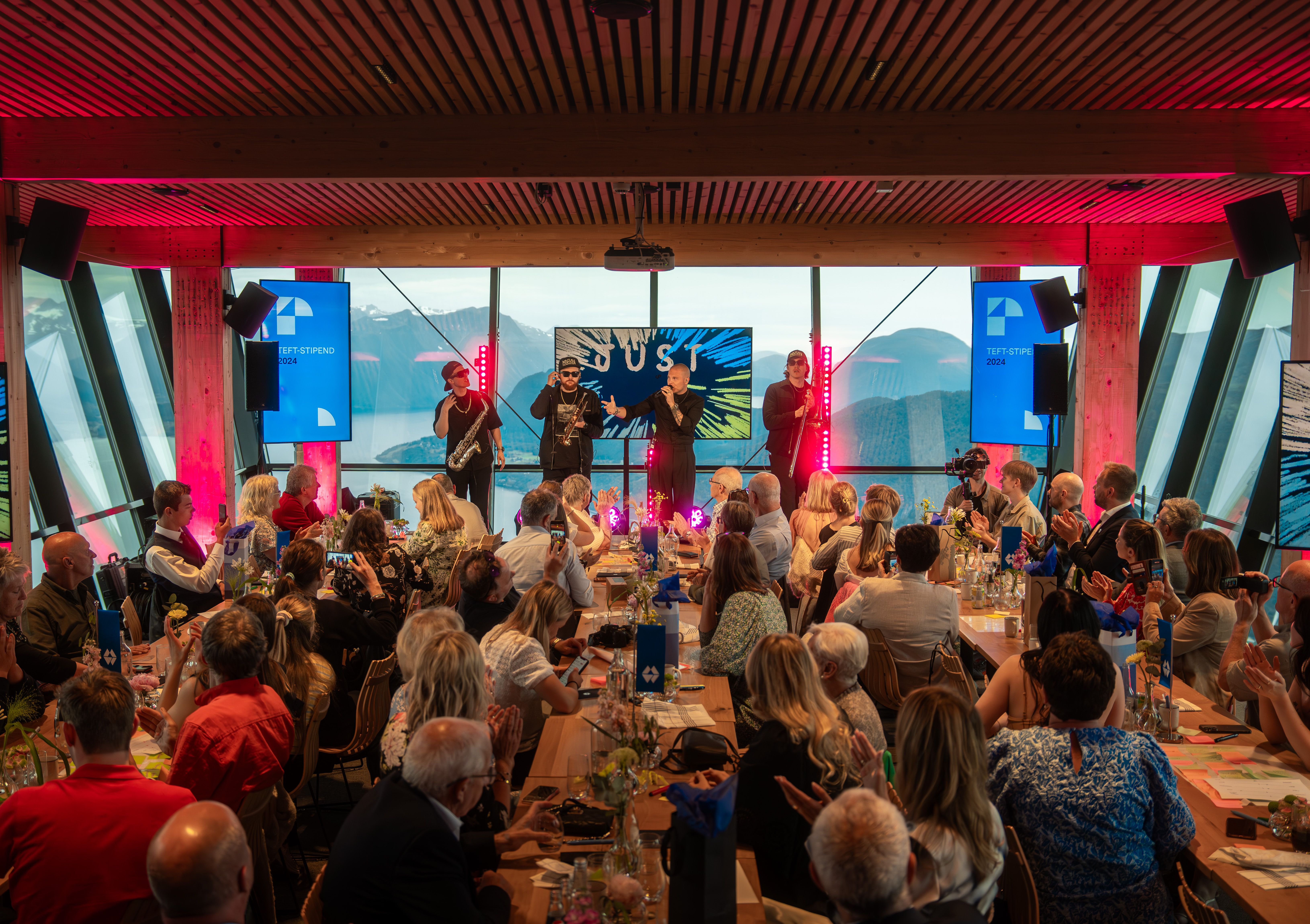 Forsamling som sitter ved langbord og ser mot 4 musikere som spiller. Rødt scenelys og landskap i bakgrunn. 