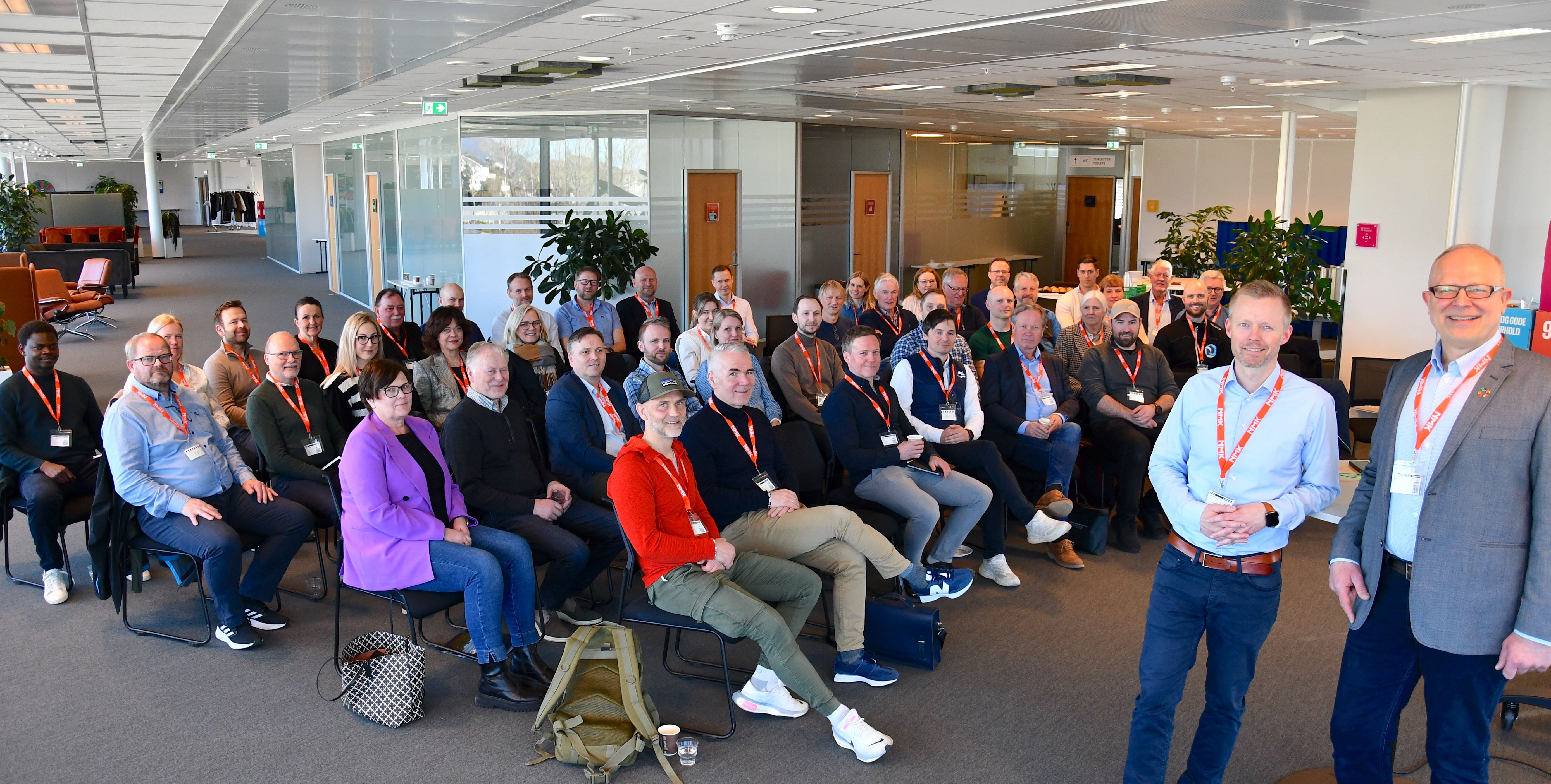 Gruppebilde av deltakere i GrønnArena Møre samlet i et møterom. Rundt femti personer sitter og står i lyse kontorlokaler, flere med oransje deltakerbånd, mens to personer står foran gruppen og smiler mot kameraet