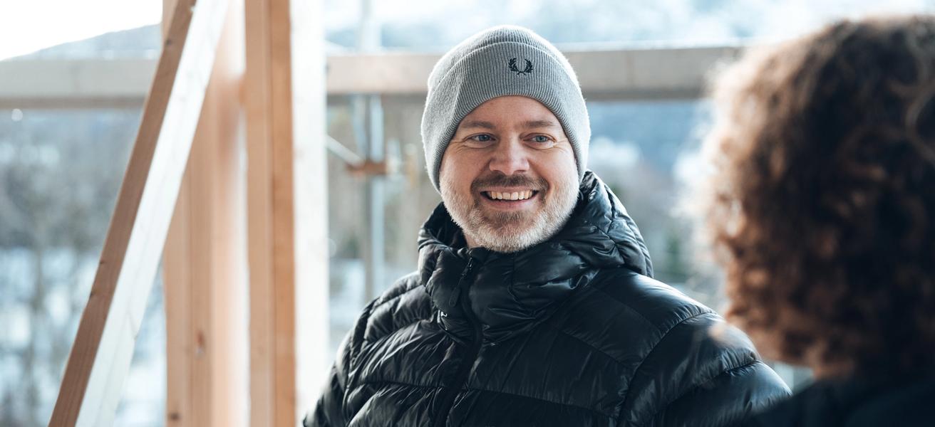 En mann med grå lue og svart jakke smiler til en person i et hus under bygging