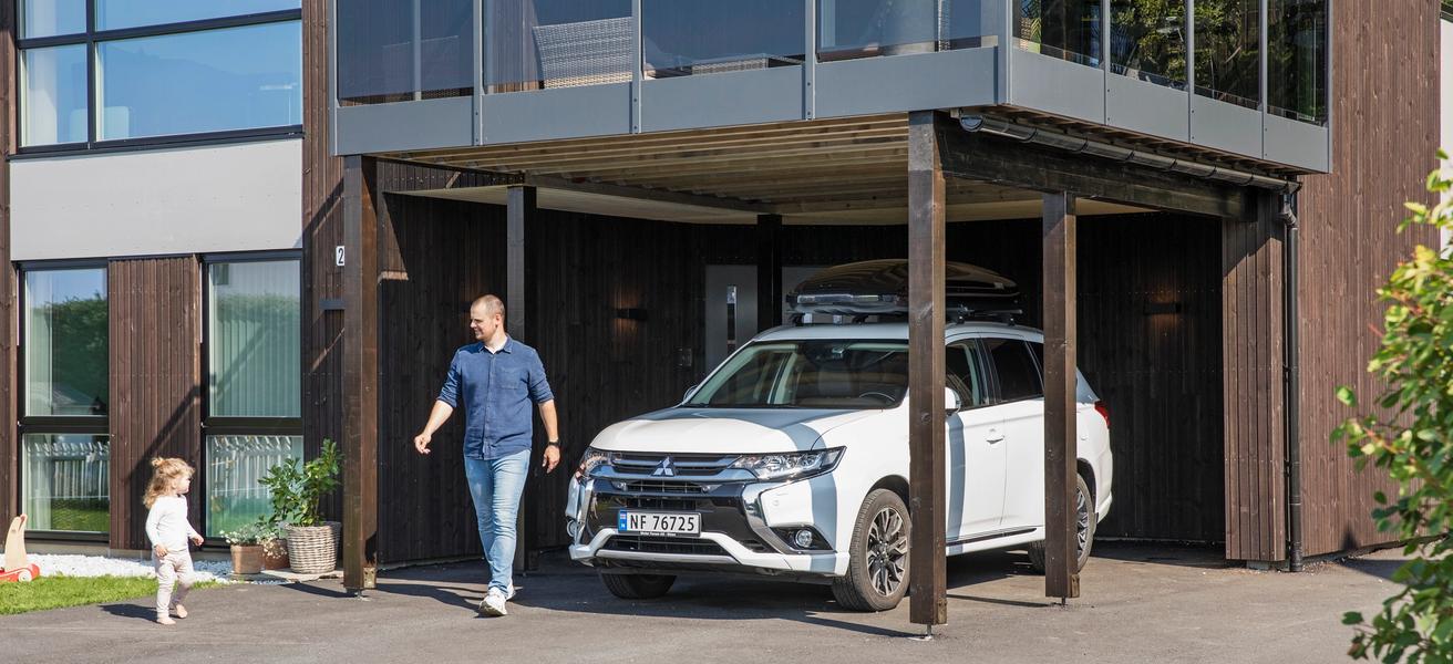 Mann og barn utenfor moderne hus, der bilen står parkert under veranda. Sol og vårlig kledde.