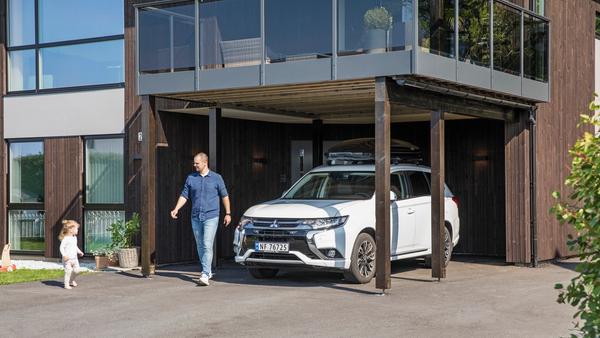 Mann og barn utenfor moderne hus, der bilen står parkert under veranda. Sol og vårlig kledde.