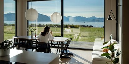Kvinne sitter ved et bord. Hun ser ut av store vinduer, der er det hav og fjell. 
