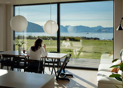 Kvinne sitter ved et bord, ser ut av store vindu ut mot havet. 
