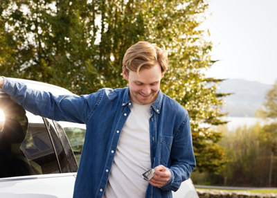 Mann står ved av siden av en bil, holder en hånd på taket på bilen. Holder bilnøklene i andre hånden, ser ned på de og smiler. 