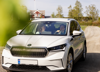 Bil kjøre ned grusvei. I passasjersetet sitter en kvinne og i førersetet sitter en mann, begge smiler. 