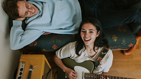 Mann ligger på en sofa og smile. En kvinne sitter på gulvet ved sofaen, spiller gitar og smiler. 