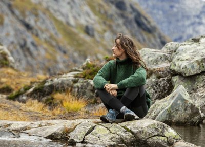 Person i grønn genser og svarte turbukser som sitter på en stein i fjellet ved vann.