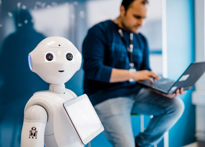 Roboten Pepper i forgrunnen, bak den sitter en mann og skriver på en bærbar PC
