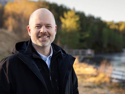 Mann står ute. I bakgrunnen er det en skog og et vann. Mannen smiler.