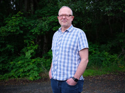 Harald fra Energiråd med rutete skjorte og briller står utendørs foran grønn skog.