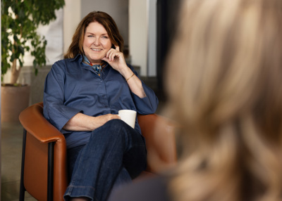 Hege Longva, daglig leder i NOA sitter smilende i lenestol med kaffekopp under en samtale i et lyst kontor eller møterom.