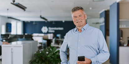 Mann i lys blå skjorte står i et moderne kontor og holder en mobiltelefon. Boliger og arbeidsplass i bakgrunnen.