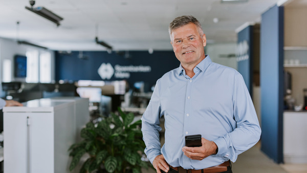 Mann i lys blå skjorte står i et moderne kontor og holder en mobiltelefon. Boliger og arbeidsplass i bakgrunnen.