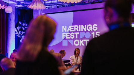 Næringsfest med scene, projeksjonslerret og publikum i et konferanselokale med lilla belysning.