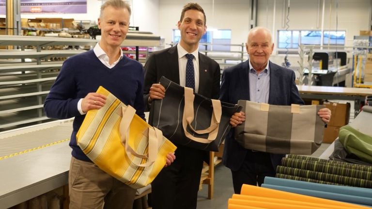 Tre personer står i produksjonslokalene hos Norsol og holder vesker laget av overskuddstekstiler fra solskjermingsproduksjonen.