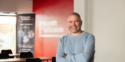 Mann med lys genser sitter på en undervisningspult, smiler til kamera. Rotevatn Trafikkskole står på en rollup i bakgrunn.