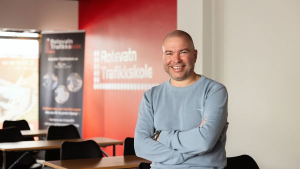Mann med lys genser sitter på en undervisningspult, smiler til kamera. Rotevatn Trafikkskole står på en rollup i bakgrunn.