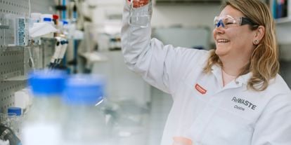 Dame i hvit frakk i et laboratorium holder oppe et glass som hun ser på. 