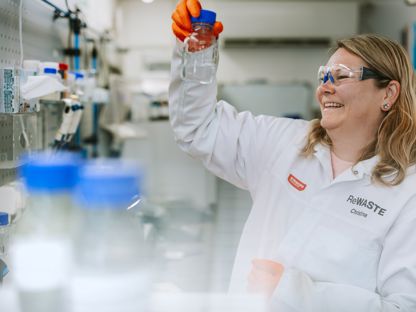 Dame i hvit frakk i et laboratorium holder oppe et glass som hun ser på. 