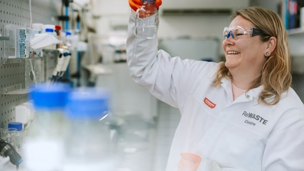 Dame i hvit frakk i et laboratorium holder oppe et glass som hun ser på. 