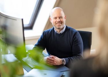 En mann som sitter på en pult med PC og har foldet hender og smiler til en person vi bare ser bakhodet til