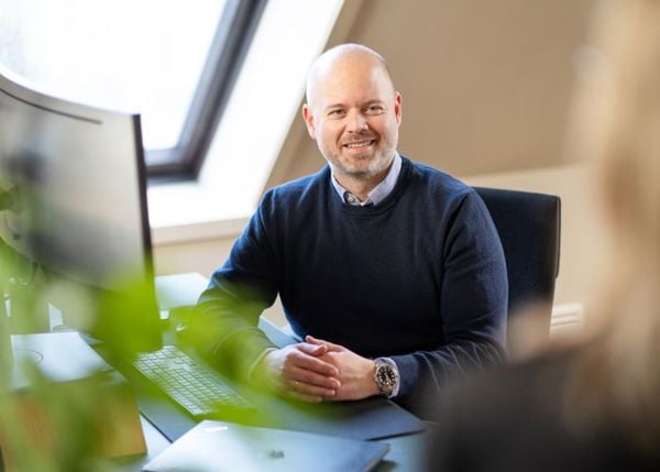 Mann som sitter med foldet hender på en pult med PC og tastatur og snakker med en person vi bare ser bakhodet på
