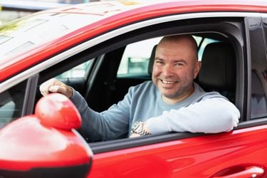 Mann med lys genser i førersetet på en rød bil. En hånd på rattet og den andre på dørkarmen.