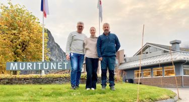 Tre personer står foran Muritunet-bygget med fjell og høstfarger i bakgrunnen. Skiltet med teksten «Muritunet» er synlig i venstre del av bildet.